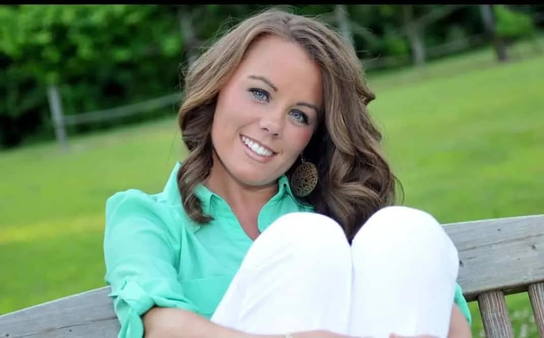 Sarah Hardison-Harris Woman with wavy brown hair wearing a green shirt and white pants, smiling while sitting on a wooden bench outdoors.
