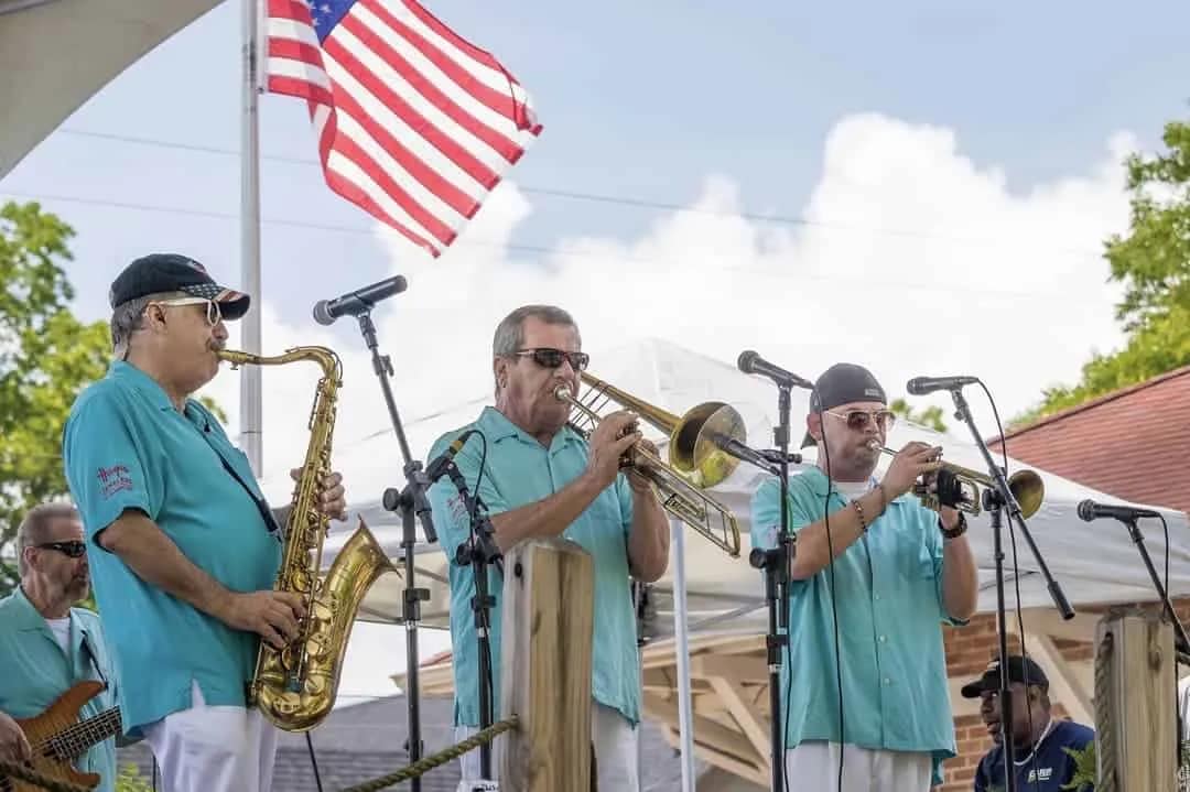 music-s Three musicians in teal shirts play saxophone, trombone, and trumpet on an outdoor stage with an American flag.