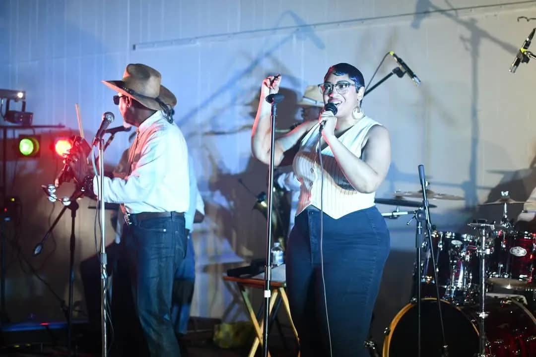 rythm-express-2 A woman sings into a microphone on stage next to a violinist; drums and instruments are in the background.