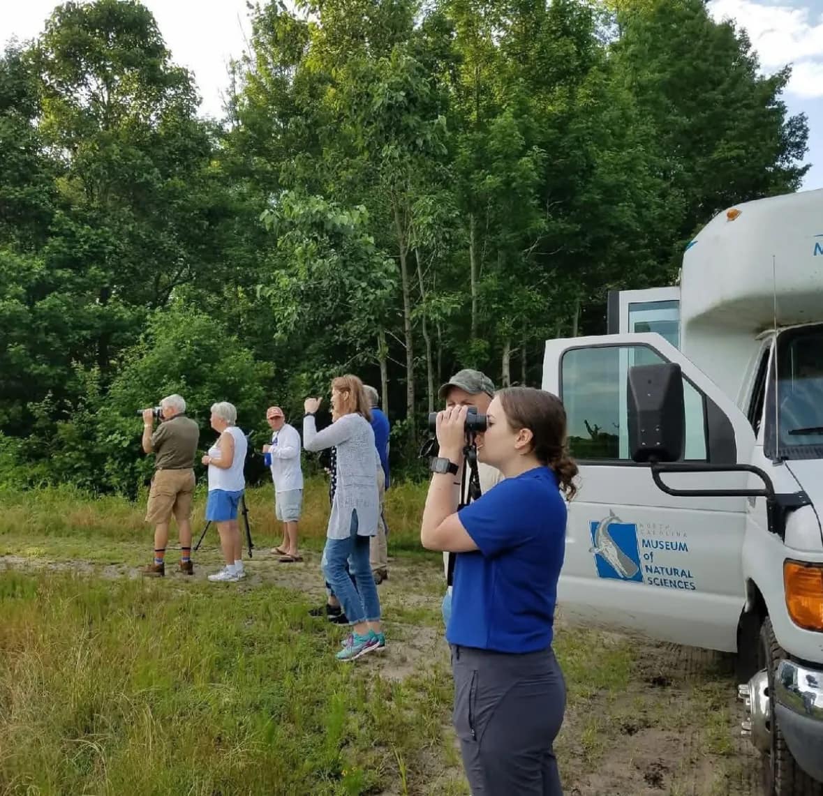 tour-2 A group of people birdwatching near a white van in a grassy area by a forest.