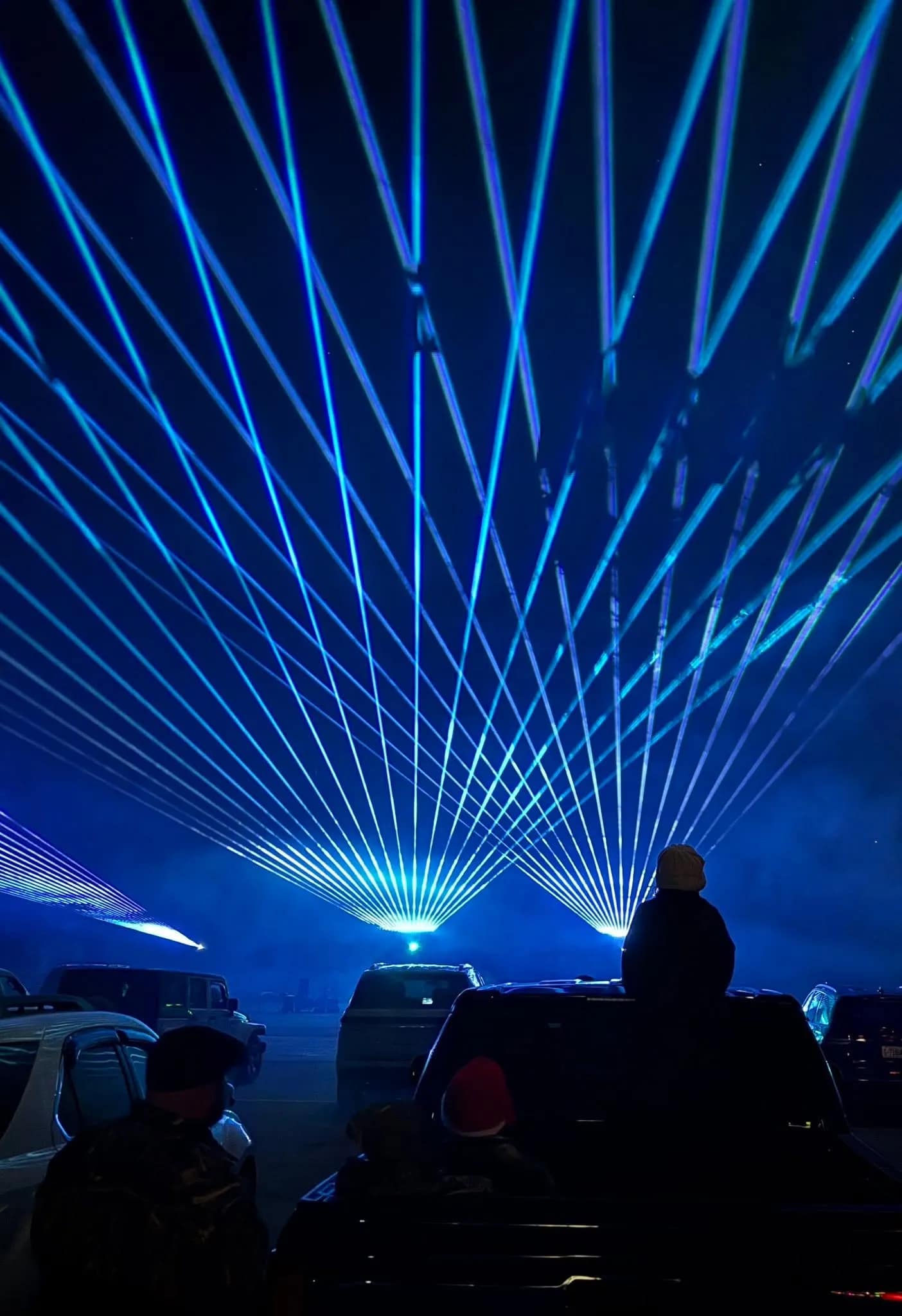 laser-1 People watch blue laser lights in the night sky from parked cars at an outdoor event.