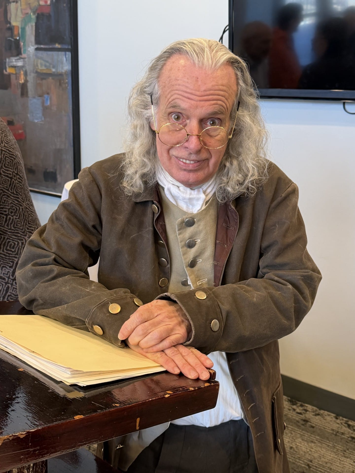 Ben-Franklin-002 An older man in colonial attire with long gray hair and glasses sits at a table with papers, smiling.