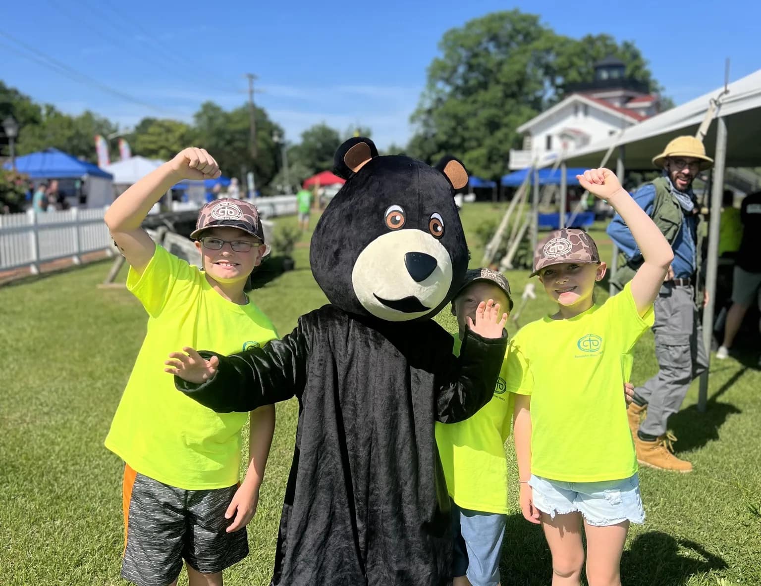 cubs-2 Three kids in yellow shirts pose with a person in a bear costume at an outdoor event on a sunny day.