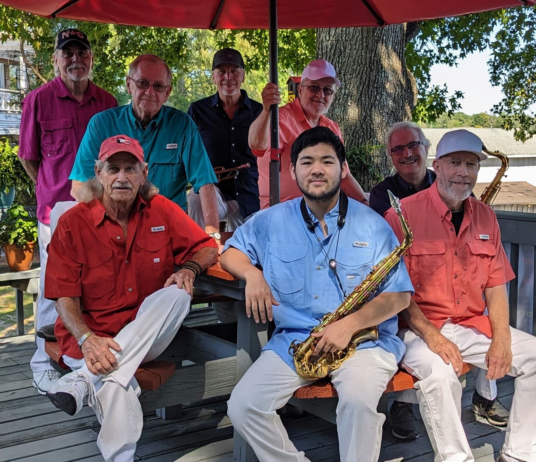 The-Patriots-Band Eight men, some holding saxophones, sit and stand on a porch under a red umbrella, smiling at the camera.