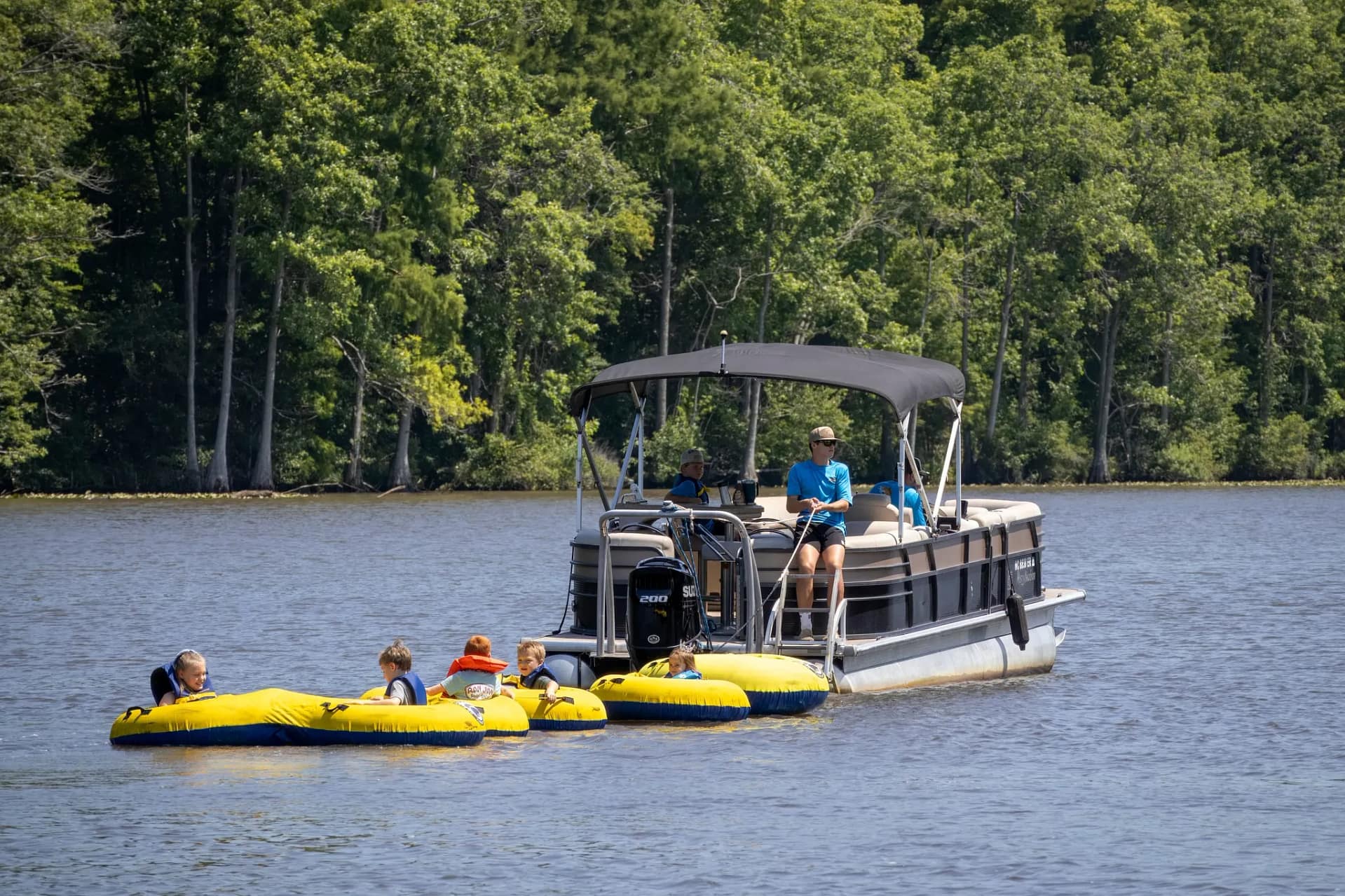 lazy-river-2 A pontoon boat pulls two yellow inflatable tubes with children on a lake surrounded by trees.