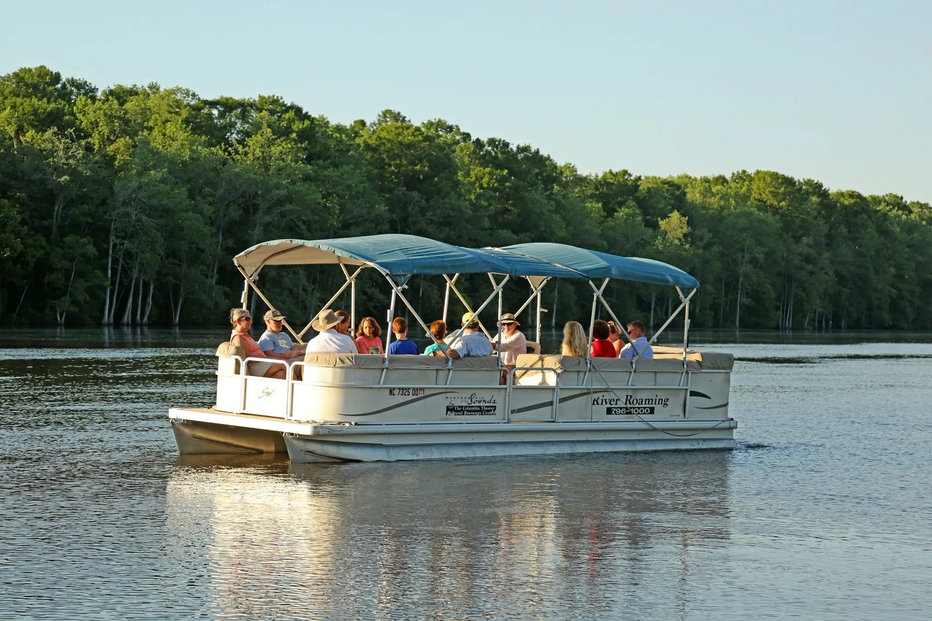 water-2 A pontoon boat with people on board cruises on a calm river with trees lining the shore in the background.