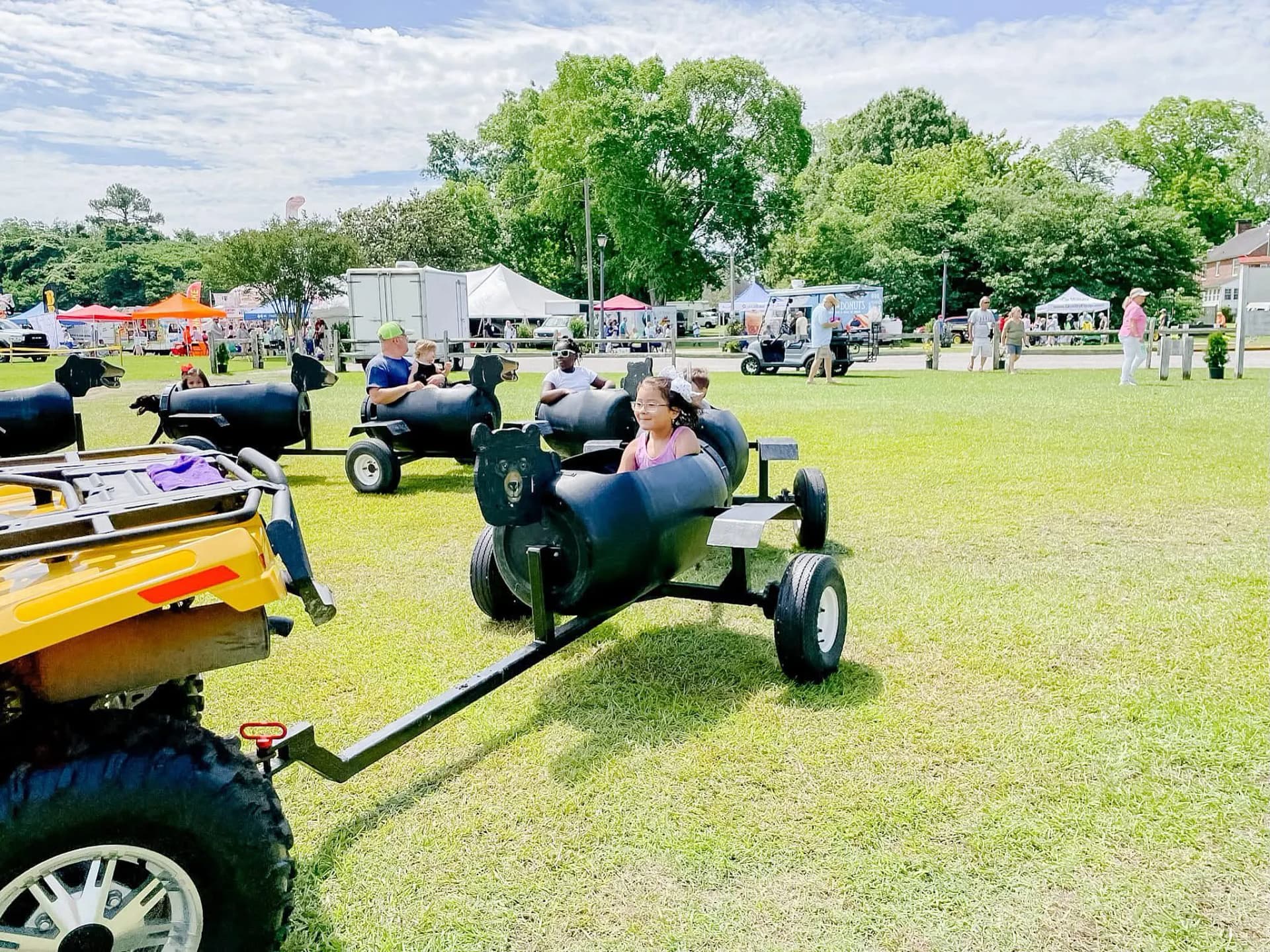 bear-train-1 Children ride in black barrel cars pulled by an ATV at an outdoor fair on a sunny day.