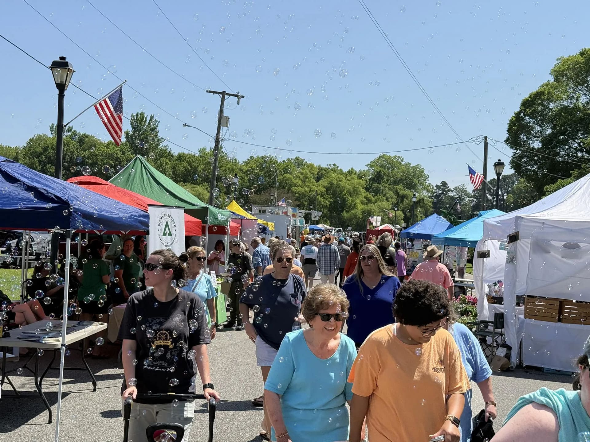 events-s People walk among colorful tents at an outdoor fair under sunny skies, with bubbles floating in the air.