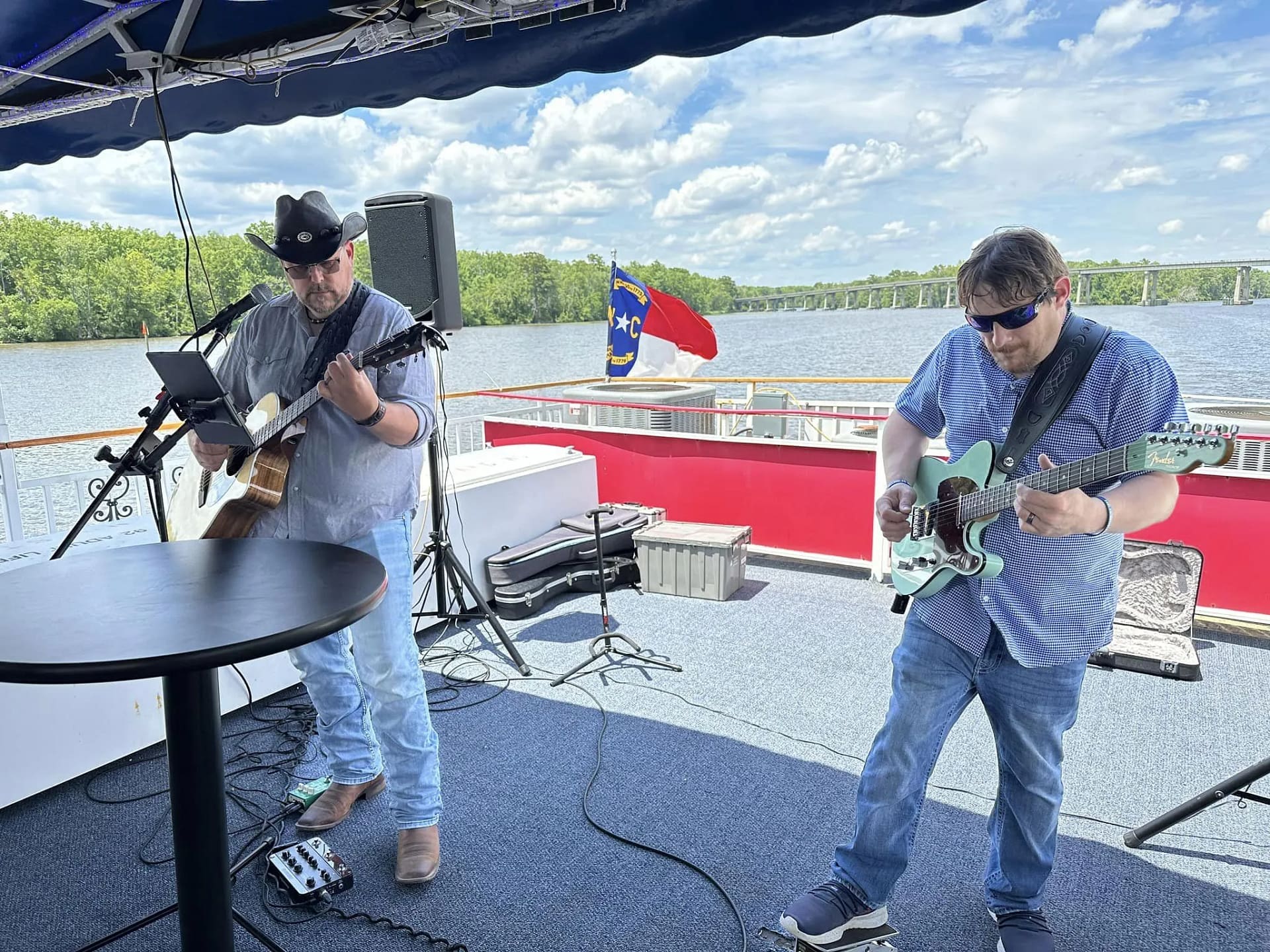 music-3 Two men play guitars on a boat deck, with a river, trees, and a flag in the background under a blue sky.