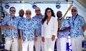 The-Main-Event-Band A diverse band of seven, including a female singer, wears matching blue floral shirts and white pants, posing for a photo outdoors with instruments.