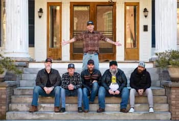 The-Daniel-Jordan-Band Six men, five seated on steps and one standing behind them, pose in front of a building with columns and glass doors.