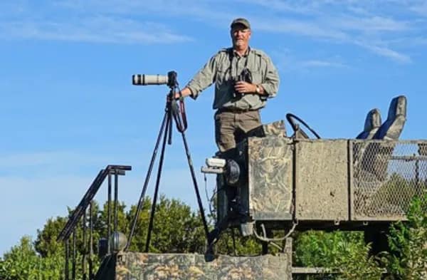 tom harrison-11 A man stands on a safari vehicle platform with a camera and tripod, surrounded by greenery and blue sky.