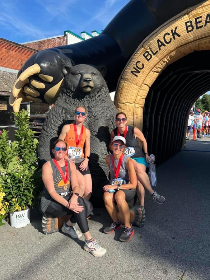 race_20368_272211_b694229d-dffa-451c-97f7-4c35b96a62a1 Four runners with medals pose by a bear statue at the NC Black Bear event entrance on a sunny day.