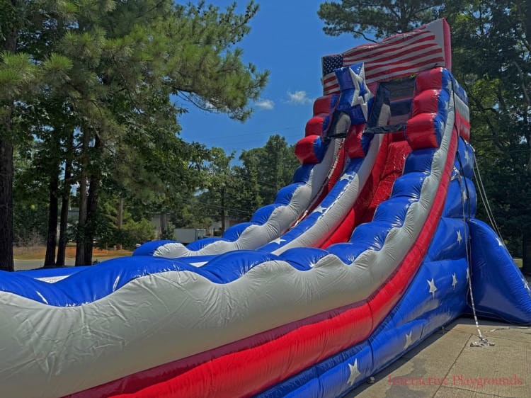 IMG_9594-side-closeup-mod-trademark-web_1751231178_big A tall inflatable slide with a stars and stripes American flag design, set outdoors near trees.