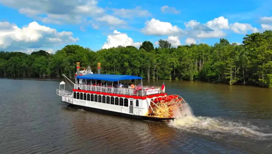 water-s A white and red riverboat with a paddle wheel cruises along a tree-lined river under a blue sky.