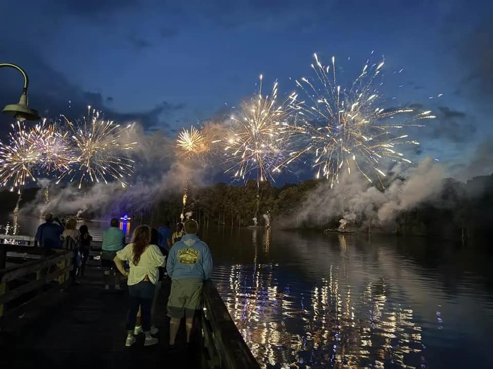 fireworks-1 People watch colorful fireworks explode over a lake at night, with reflections shining on the water.