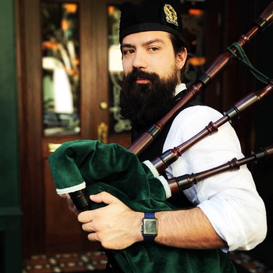 Bagpipes-002 A man with a beard plays the bagpipes, wearing a hat and a smart vest, standing by wooden doors.
