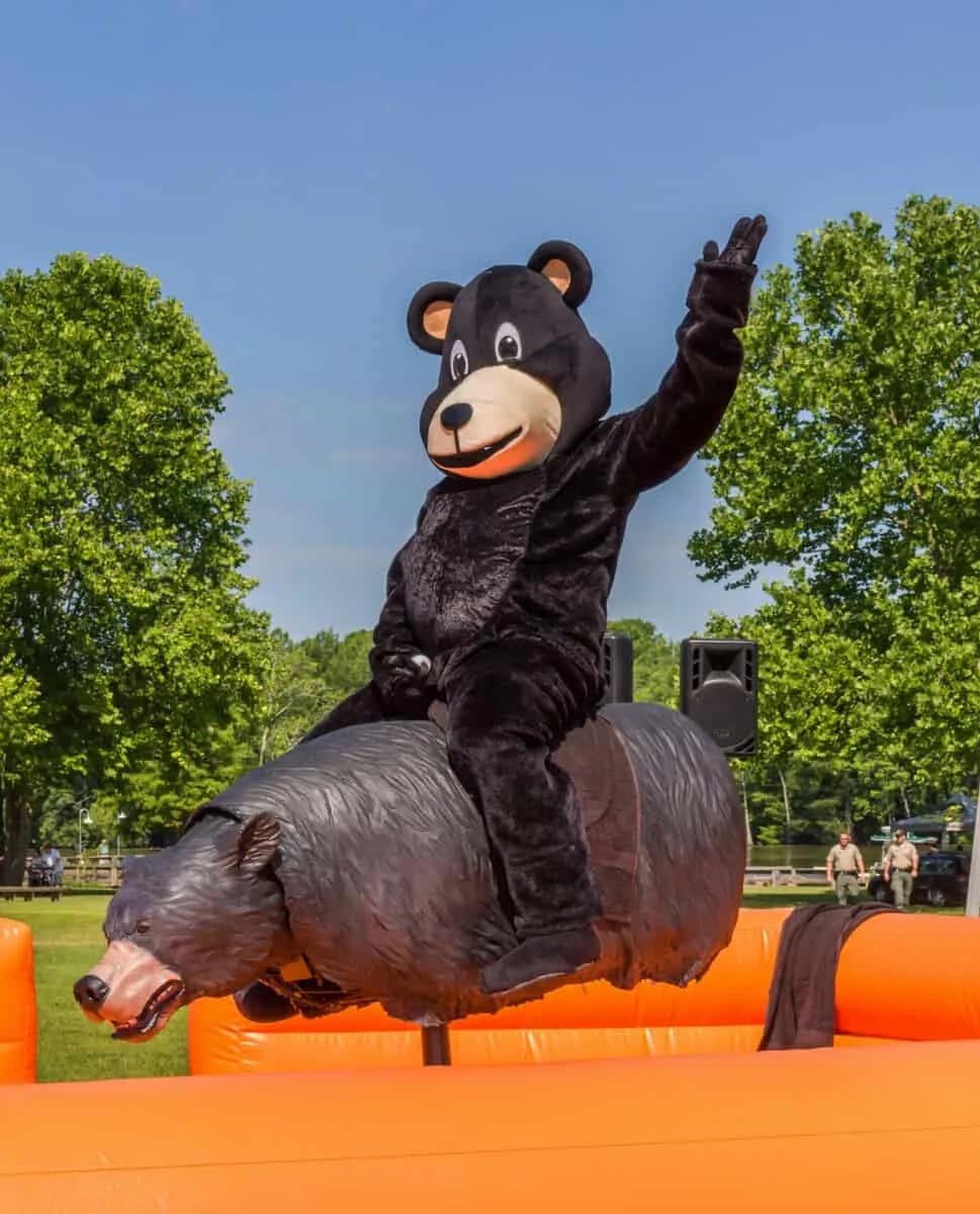 why-choose-1 Person in a black bear costume rides a mechanical bear at an outdoor event with trees in the background.