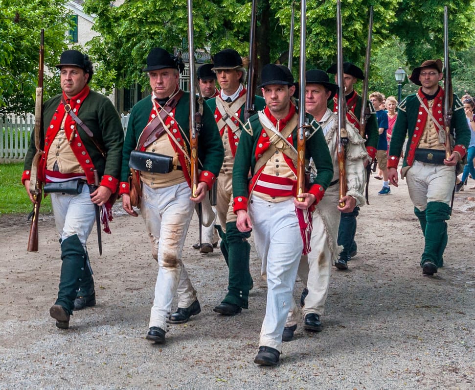 Rev-War-Soldiers-dreamstime_m_105995619 A group of reenactors in Revolutionary War-era uniforms march outdoors on a gravel path.